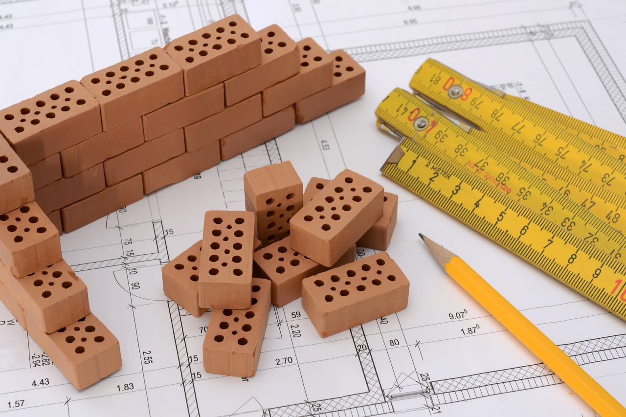 Symbolbild Kleine Ziegelsteine, ein Zollstock und Bleistift liegen auf dem Bauplan für ein Haus