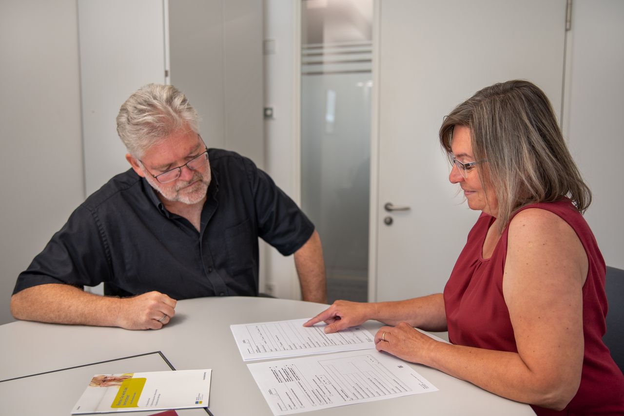 Eine Mitarbeiterin der Gemeinde sitzt mit einem älteren Mann am Schreibtisch, deutet auf Papiere der Rentenversicherung und berät den aufmerksamen Klienten.