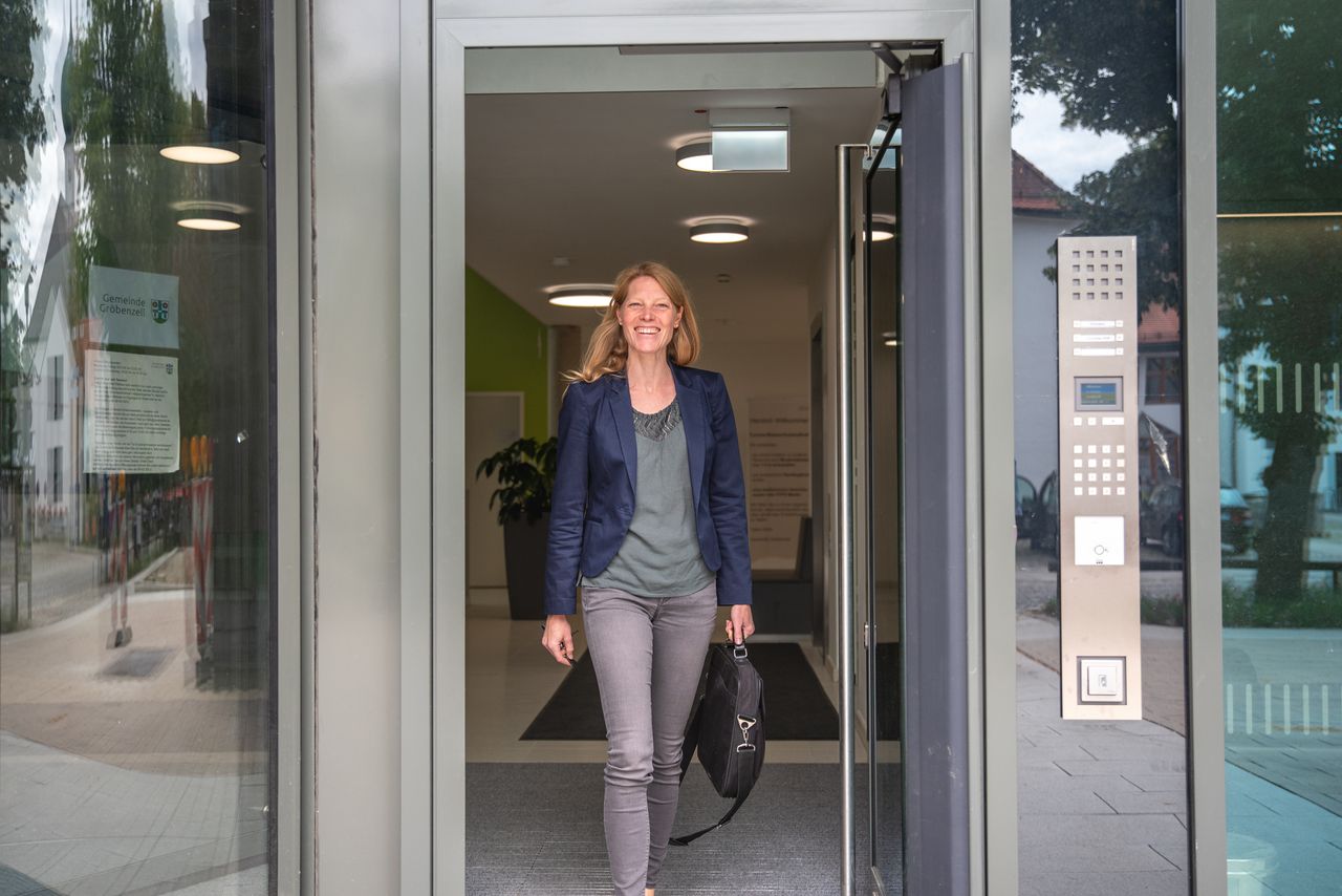 Lachende Mitarbeiterin im Blazer verlässt mit Laptoptasche in der Hand das Rathaus durch den gläsernen Haupteingang.