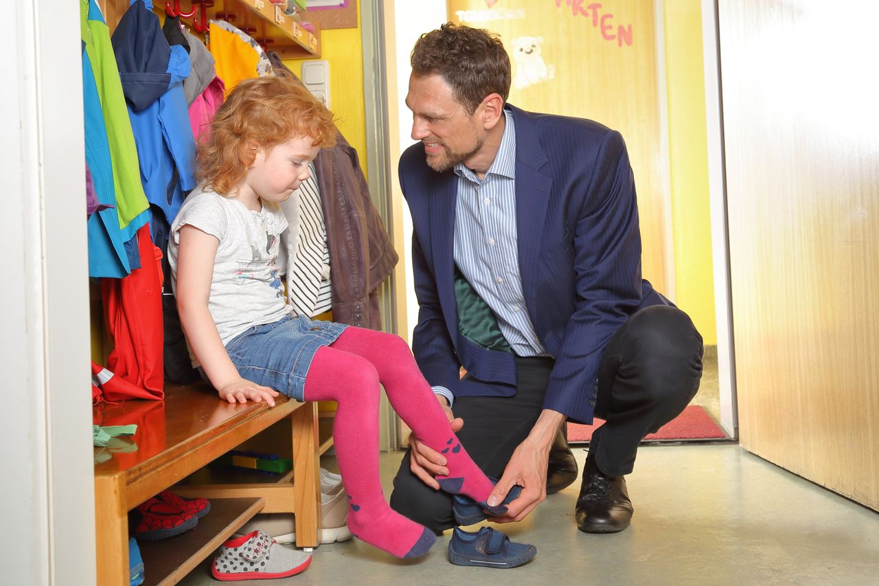 Vater in einem Business Casual Outfit zieht Tochter im Kindergarten die Hausschuhe an