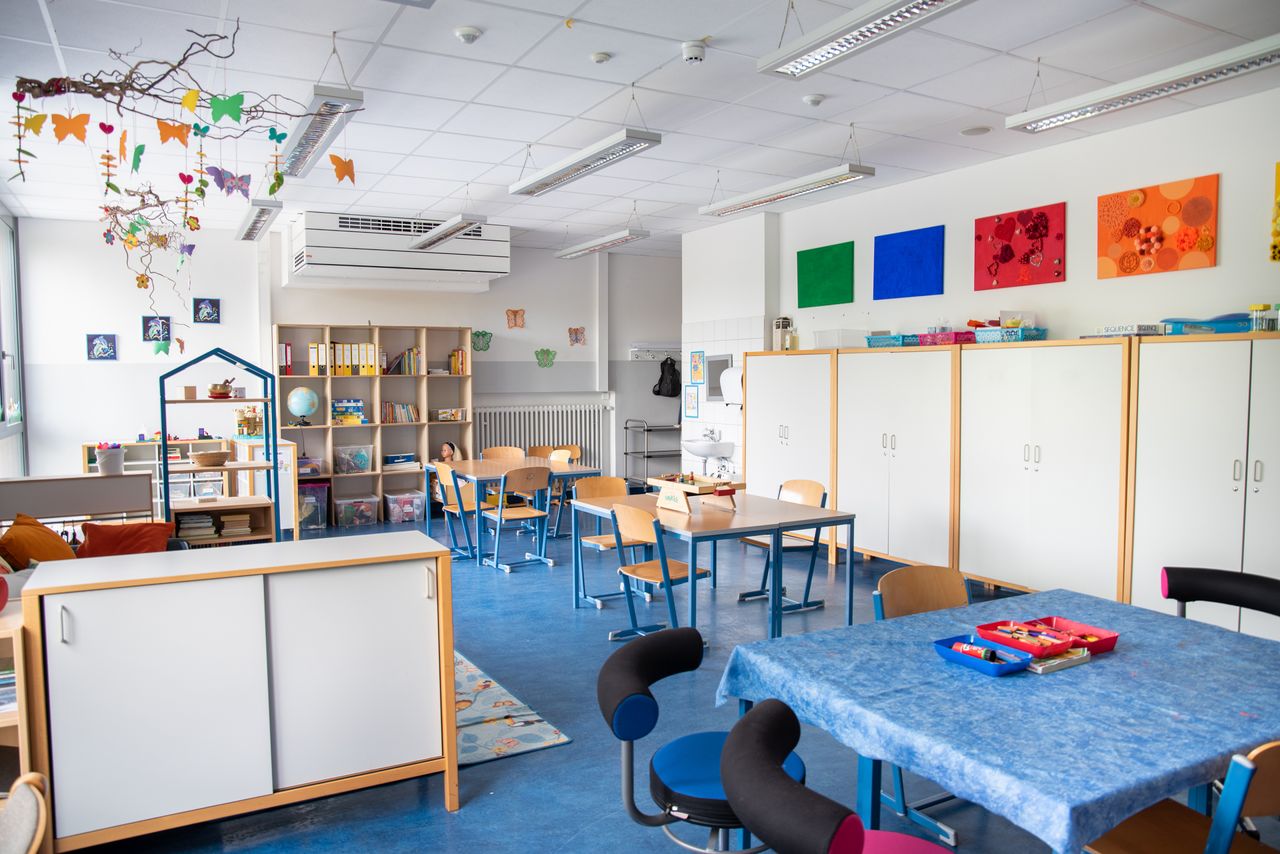 Mittagsbetreuung in der Ährenfeldschule. In der Raummitte Gruppentische mit Stühlen. Links zwei Spielecken mit Spielzeug und Regalen.