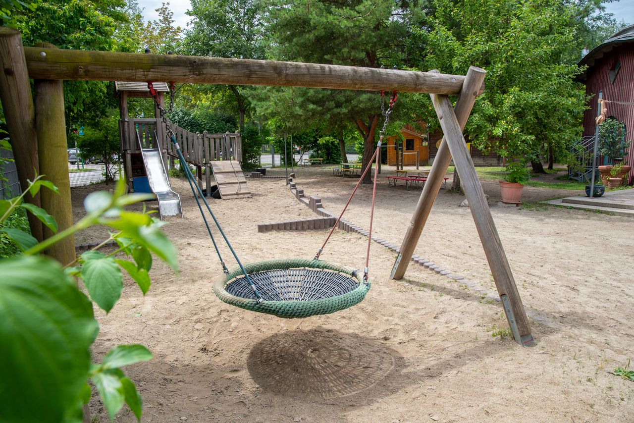 Nestschaukel im Garten des Kindergartens, im Hintergrund ein Spielturm mit Rutsche, rundherum ein Sandkasten, im Hintergrund Sitzbänke und eine Terrasse