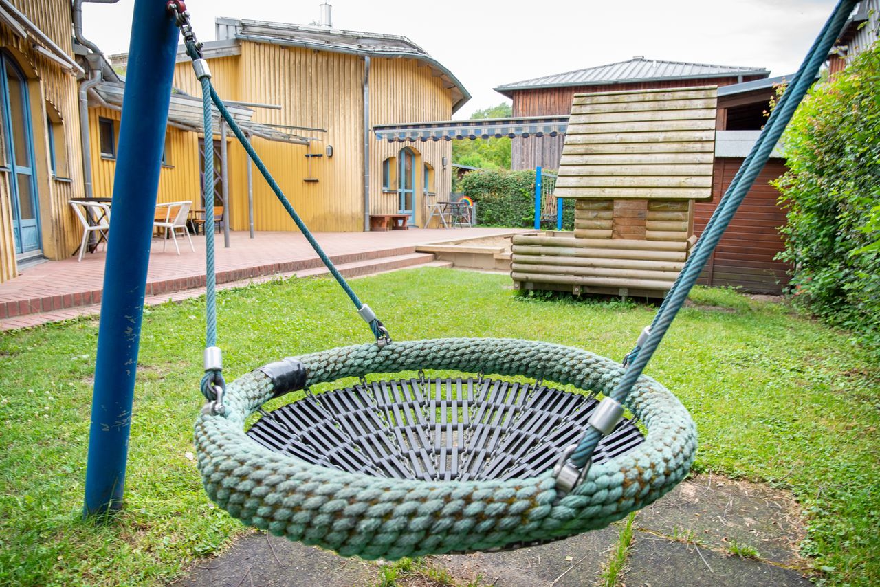 Nestschaukel im Garten der Kinderkrippe, im Hintergrund ein Spielhaus und eine Terrasse mit Stühlen und Bänken