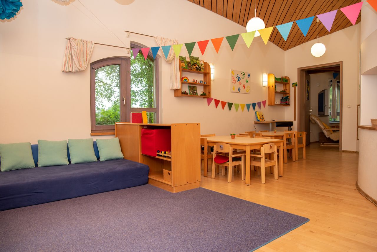 Ein Gruppenraum in der Kinderkrippe Unterm Regenbogen mit einer Kuschelecke, einem Teppich, Stühlen und Gruppentischen