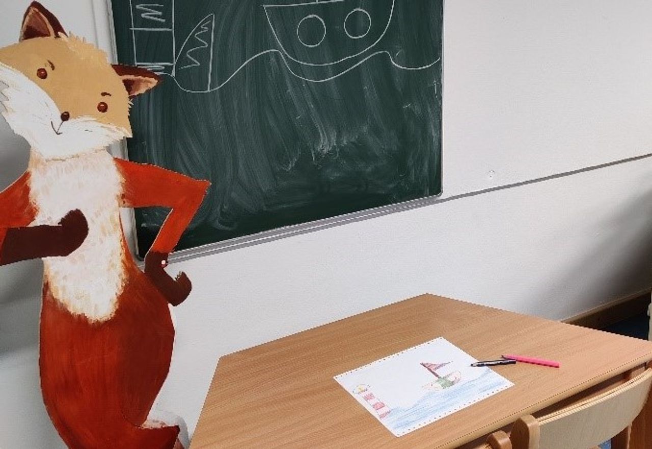 Ein Fuchsaufsteller, vor ihm ist ein Tisch und hinter ihm ist eine Tafel. Auf dem Tisch ist ein gemaltes Bild.