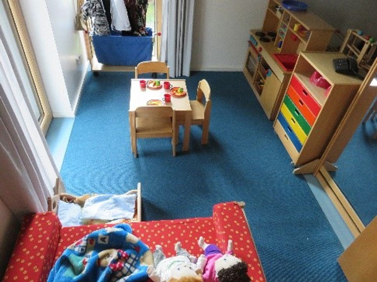 Ein kleiner Raum von oben mit blauem Teppich, Tisch und drei Stühlen in der Mitte. An der Wand steht eine Spielküche.