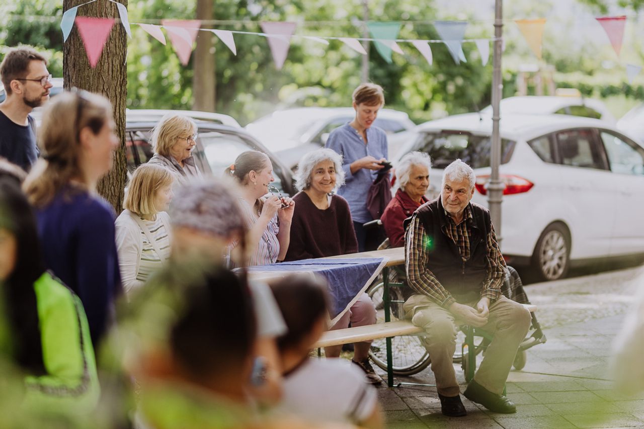 Jung und Alt aus der Nachbarschaft versammeln sich an einer Bierzeltgarnitur, die auf dem Bürgersteig steht,  im Hintergrund parkende Autos.
