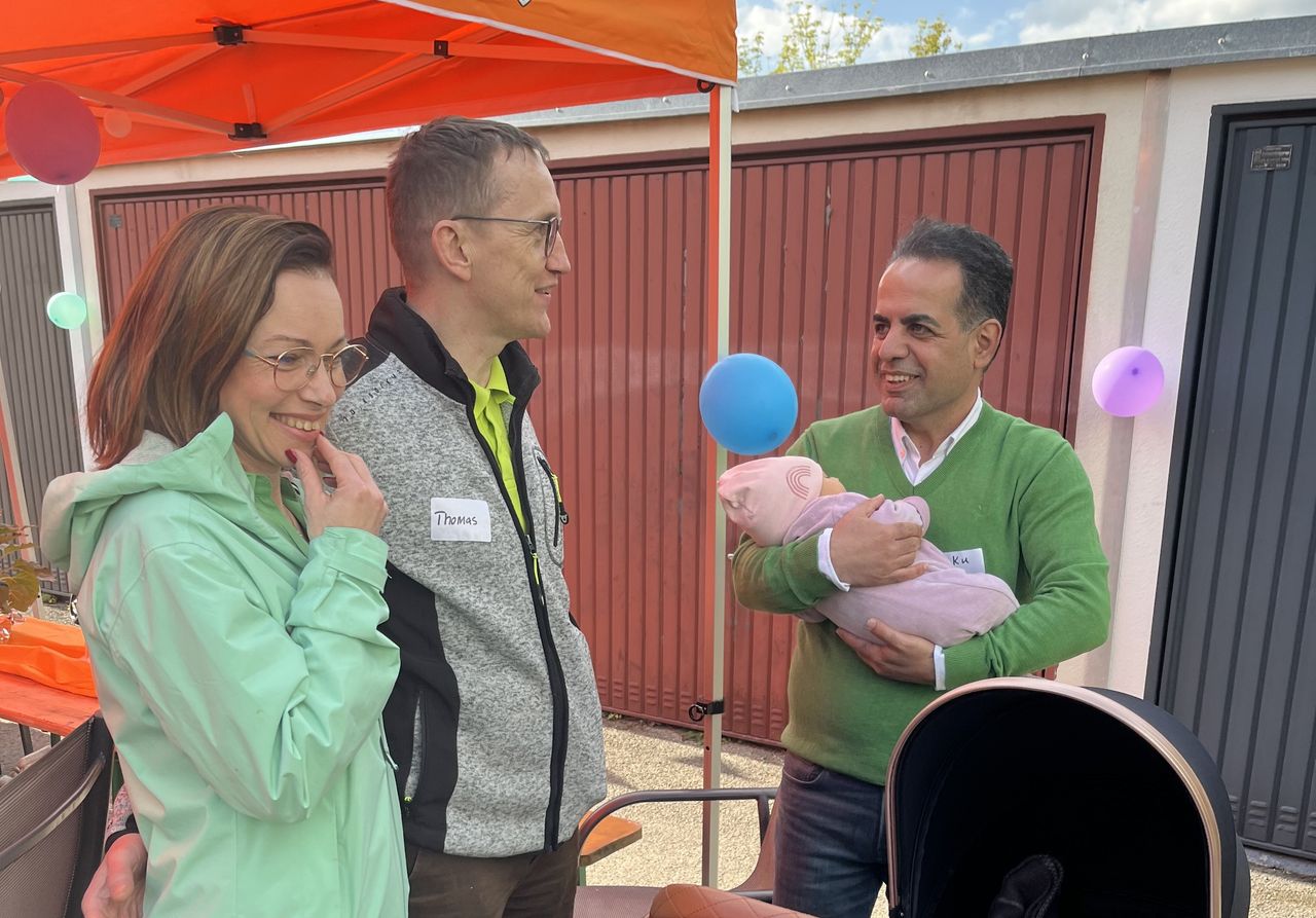 Nachbarn unterhalten sich angeregt beim Garagenfest unter einem mit Ballons  geschmückten Pavillon, ein Vater hält dabei sein Baby auf dem Arm