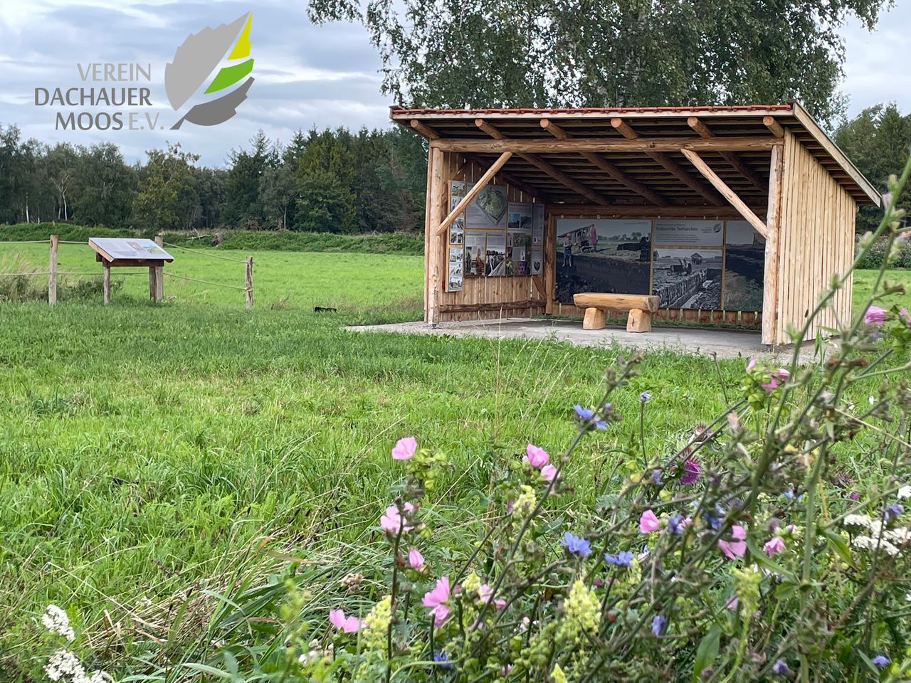 Foto der Torfhütte, davor blühende Blumen, viel Wiese.
