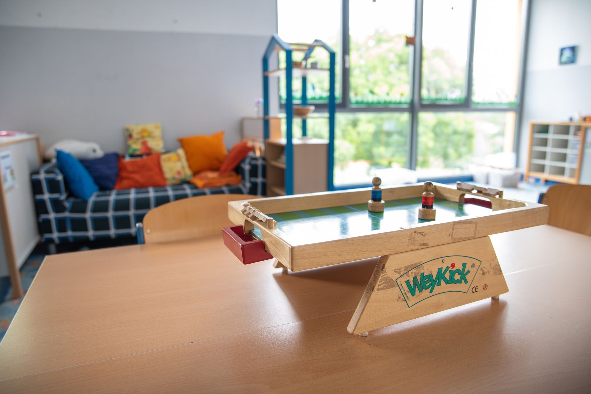 Ein kleiner Tischkicker steht auf einem Gruppentisch in der Mittagsbetreuung der Ährenfeldschule, im Hintergrund eine Spielecke, Regale und eine Couch mit Kissen darauf