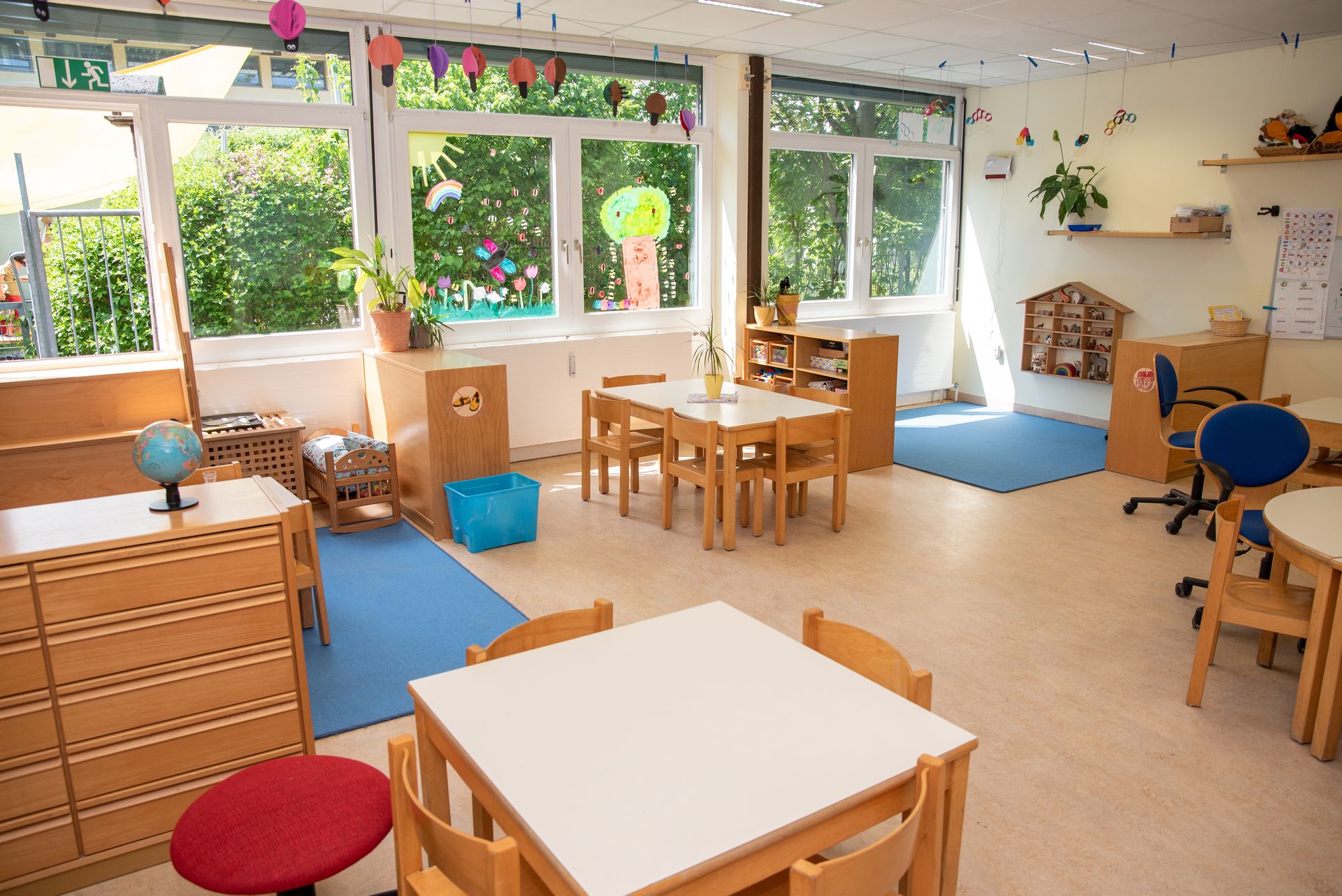 Gruppenraum im Kindergarten Wichtelhaus mit eckigen Gruppentischen und Stühlen rundherum sowie blauen Teppichen am Boden