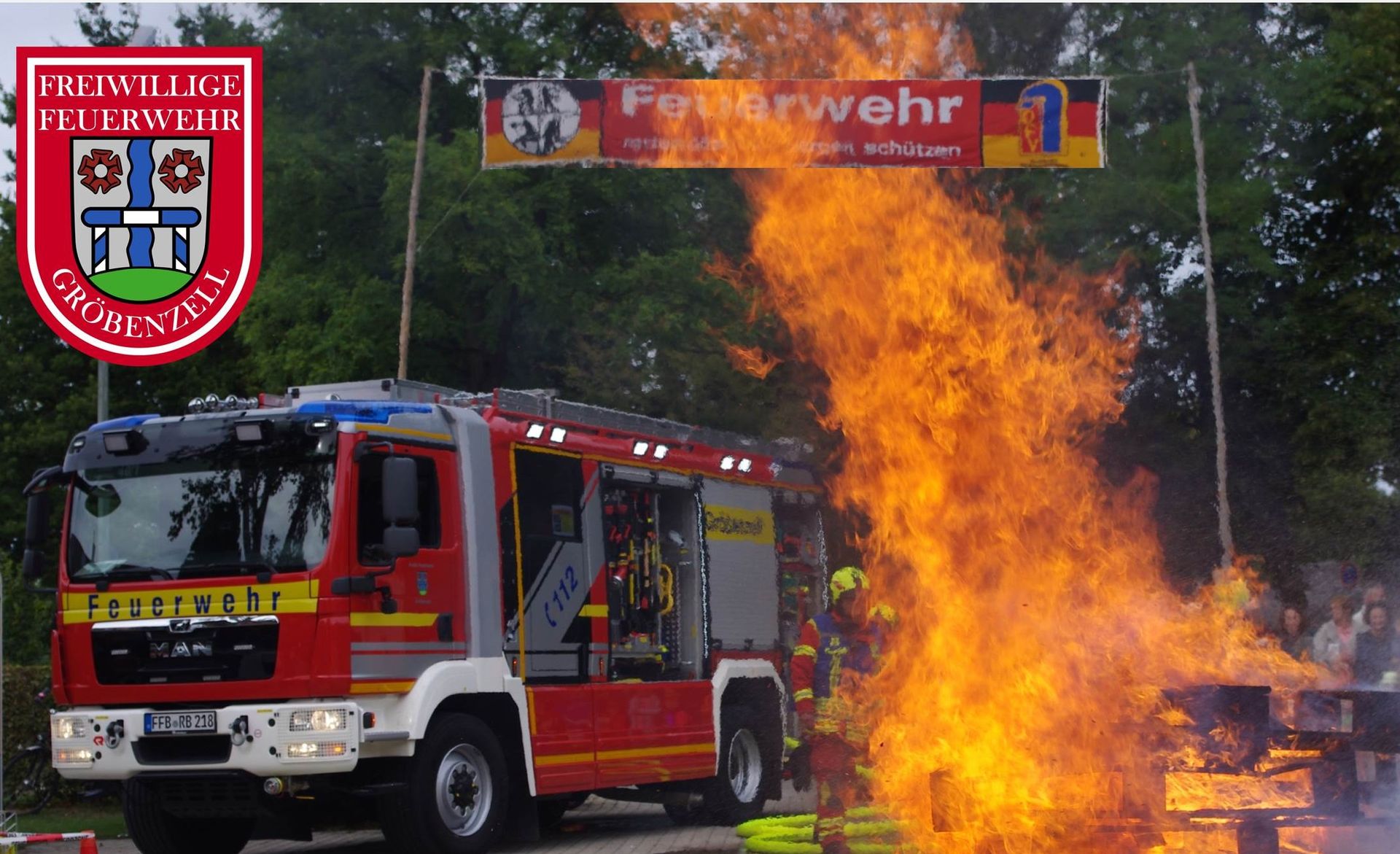 Feuerwehrauto im Hintergrund, davor Flammen. Löschübung vor Publikum