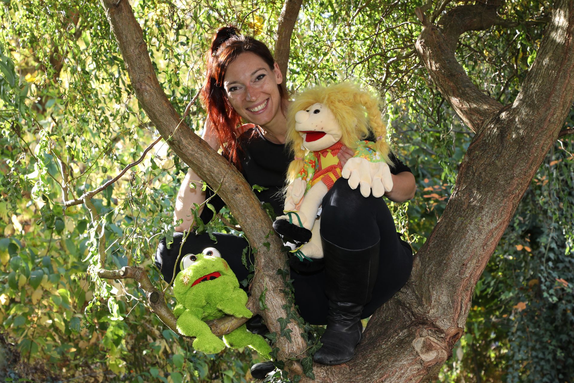 Prinzessin Alleswill und der kluge Frosch Eine rothaarige Frau hockt auf einem Baum. Auf einem Ast unte4r ihr sitzt ein grüner Plüschfrosch. Auf einem Knie sitzt eine Mädchen-Handpuppe mit wuscheligen gelben Haaren.