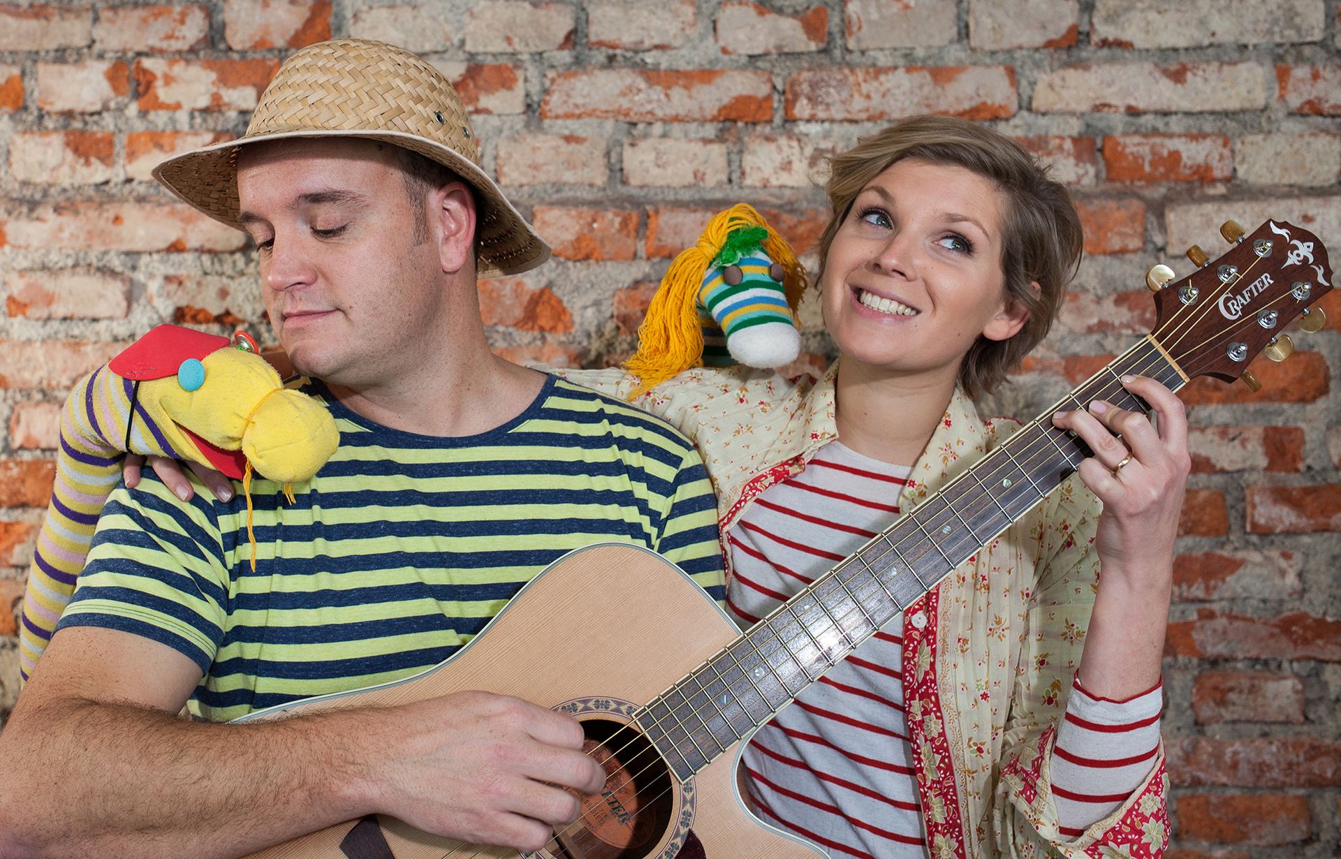 Mit Zick & Zack auf Geschichtenreise – das Mitspinntheater für Kinder und zwei Ringelsocken Ein Mann mit Strohhut hält eine Gitarre und spielt darauf gemeinsam mit einer lachenden Frau, die rechts neben ihm steht. Beide haben bunte Sockenpuppen auf den Schultern.