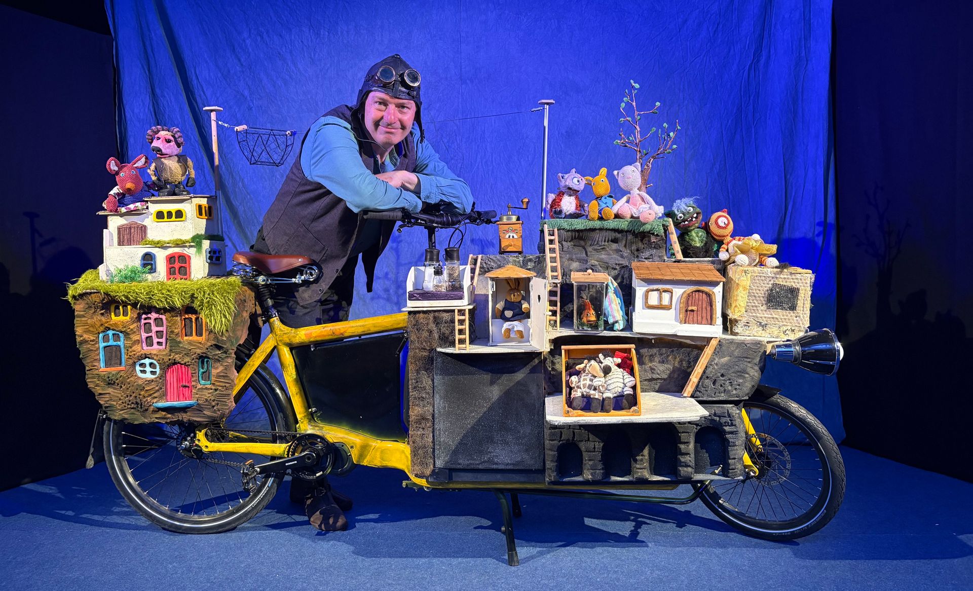 Kindertheater: Rollende Stadt Mann mit Fliegerkappe lehnt sich auf den Lenker am zum Puppentheater umgebauten Fahrrad.