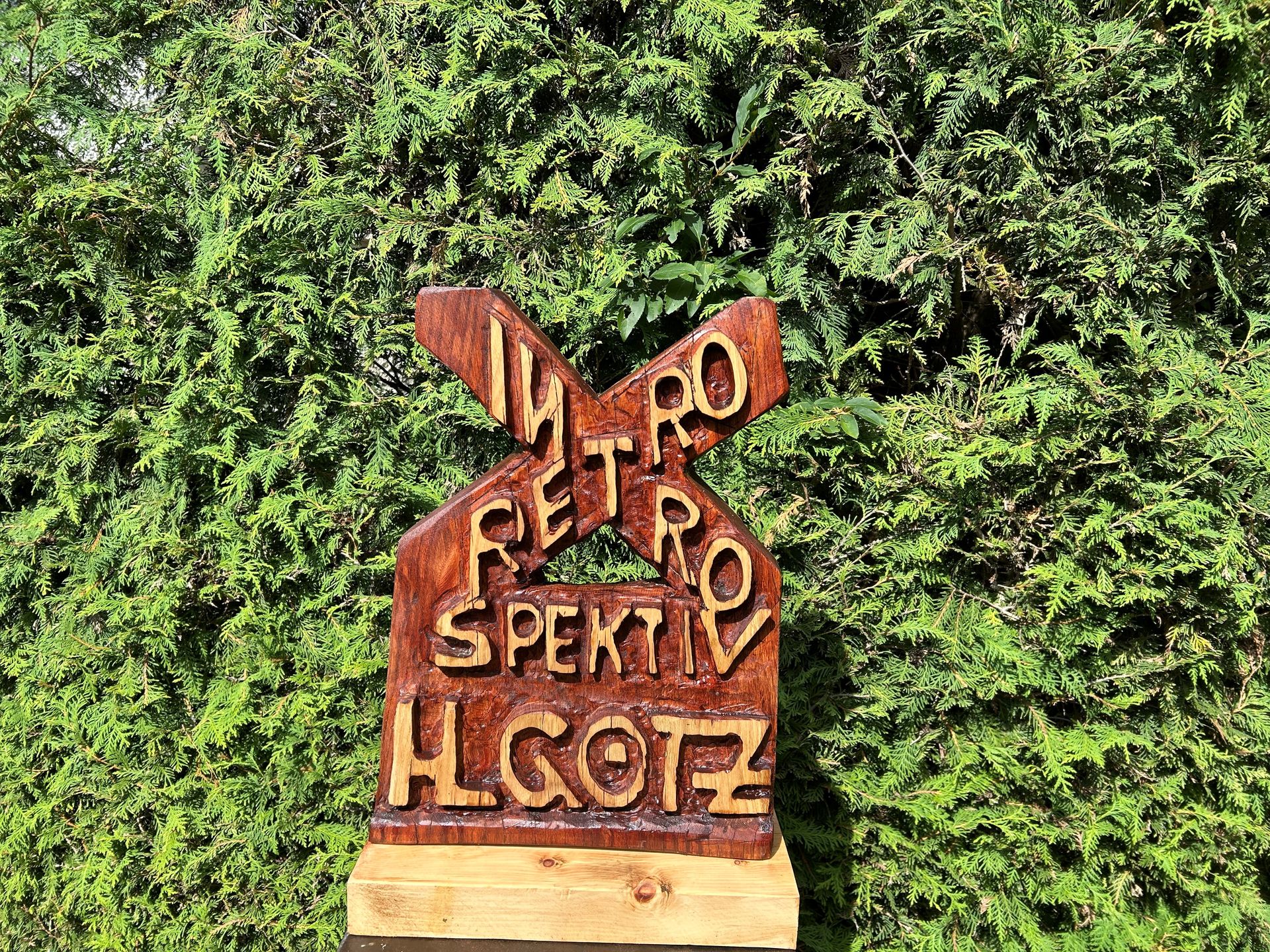 Ausstellung „Retrospektiv / Introspektiv“ von Hannes L. Götz Holzskulptur zeigt Logo der Ausstellung Retrospektiv - introspektiv. "Retro" "Intro" als X überkreuzend und darunter "spektiv". Ganz unten H. Götz
