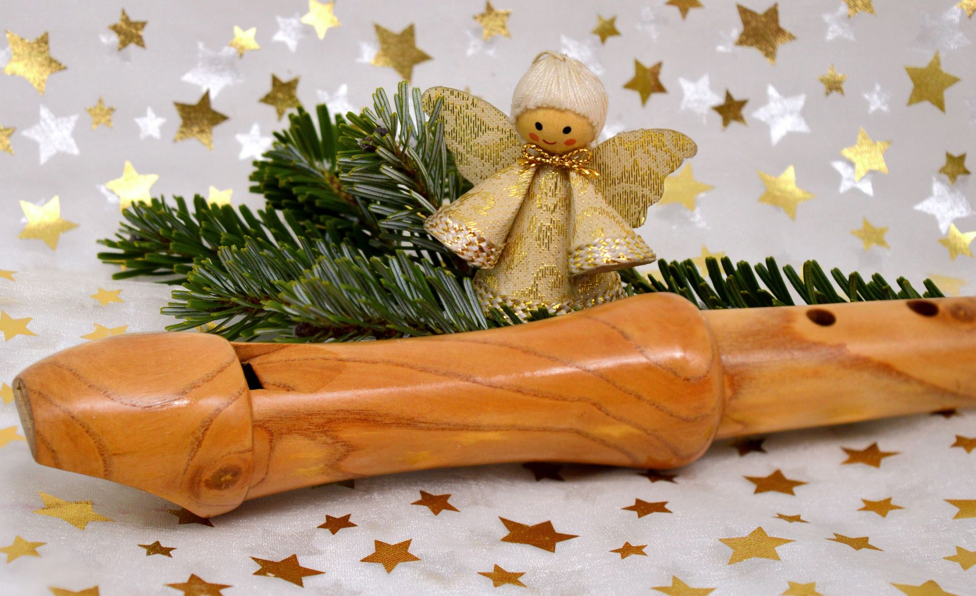 Ein Sopran-Blockflöte liegt auf einem weißen Untergrund mit goldenen und silbernen Sternen. Hinter ihrsteht ein kleiner goldener Engel umgeben von Tannenzweigen.