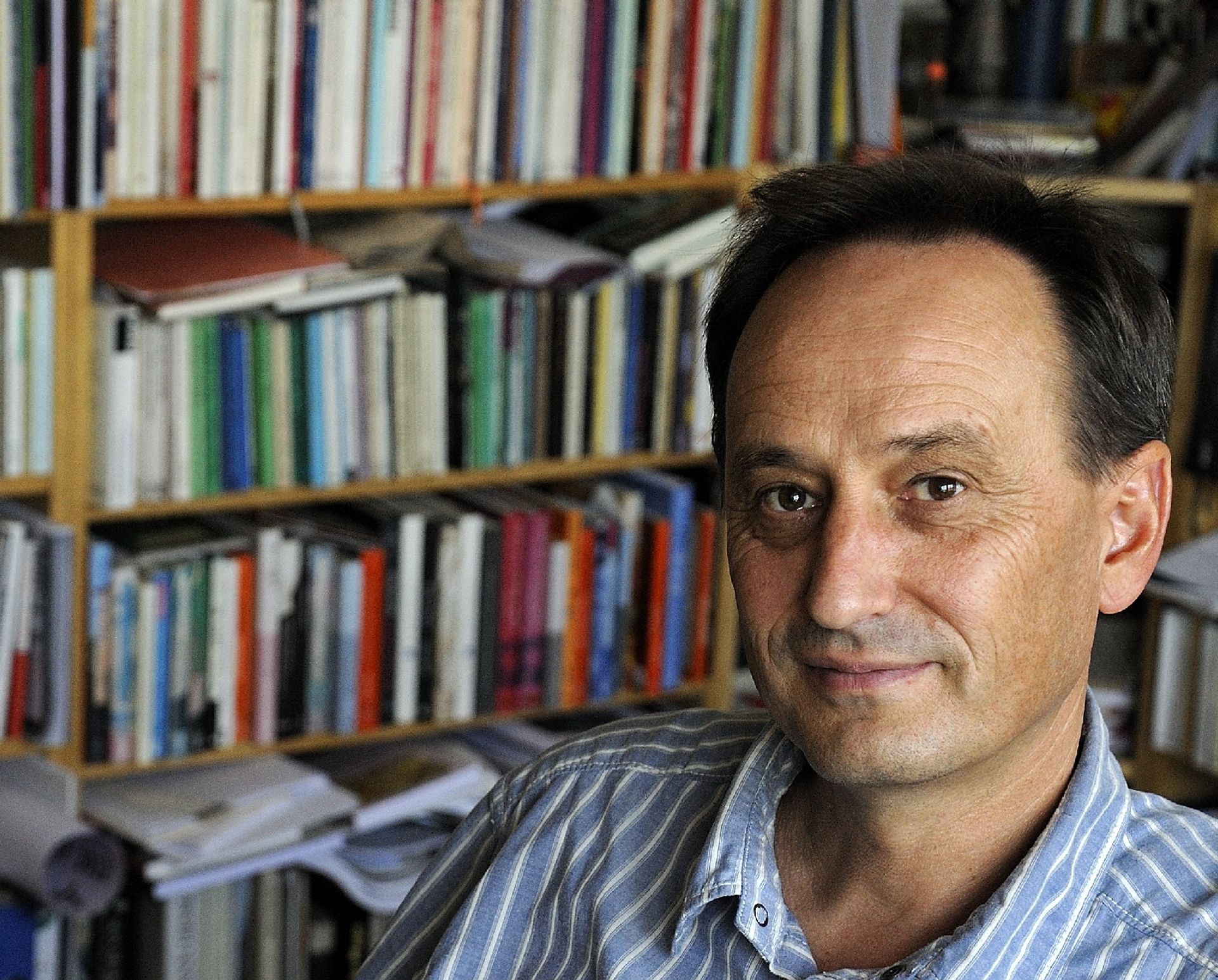 Alois Prinz sitzt, bekleidet mit einem blau-weiß-gestreiften Hemd mit offenem Kragen, vor einem bunten Bücherregal