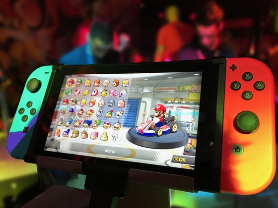 Eine Nintendo Switch Konsole mit je einem grünen und orangenem Joy Con an den Seiten zeigt auf dem Bildschirm das Spiel Mario Kart.