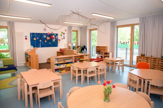Ein Gruppenraum im Kindergarten Flohkiste mit eckigen und runden Tischen und Stühlen, im Hintergrund eine Spielecke, links eine Leseecke