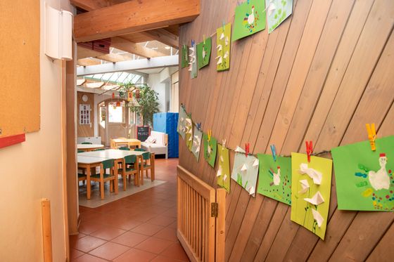 Gang im Innenbereich des Kindergartens Spatzennest. Am Ende des Gangs stehen Gruppentische mit Stühlen rundherum, an der Holzwand hängen gebastelte Bilder der Kinder.