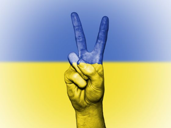 Ukrainische Flagge, im Vordergrund eine Männerhand, die ein Peace-Zeichen zeigt