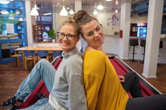 Steffi und Anna vom JBS-Team lehnen lächelnd Rücken an Rücken aneinander