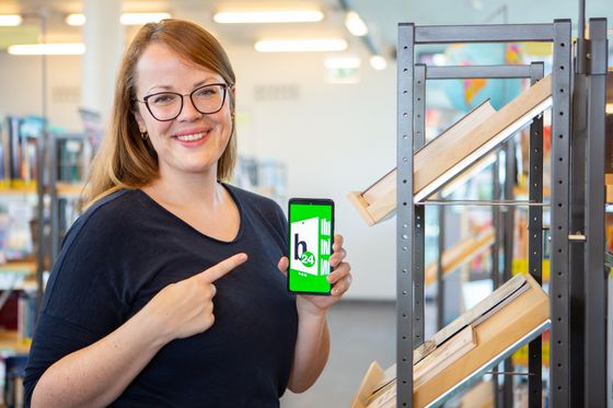 Lächelnde Frau steht am Büchereiregal und deutet auf ihr Handy