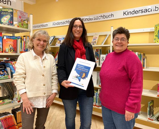 Mitarbeiterinnen der Gemeindebücherei präsentieren die Urkunde vor Bücherregalen stehend.