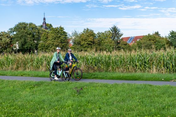 Zwei Personen mit Helmen auf dem Fahrrad, im Hintergrund ein Maisfeld, Dächer und ein Kirchturm