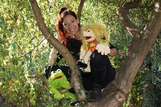 Prinzessin Alleswill und der kluge Frosch Eine rothaarige Frau hockt auf einem Baum. Auf einem Ast unte4r ihr sitzt ein grüner Plüschfrosch. Auf einem Knie sitzt eine Mädchen-Handpuppe mit wuscheligen gelben Haaren.