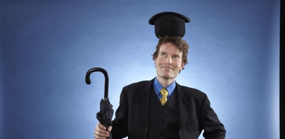 Zauberer René im schwarzen Anzug mit blauem Hemd und gelber Krawatte balanciert einen schwarzen Hut verkehrt herum auf seinem Kopf und hält einen geschlossenen Regenschirm in seiner rechten Hand