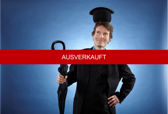 Zauberer René im schwarzen Anzug mit blauem Hemd und gelber Krawatte balanciert einen schwarzen Hut verkehrt herum auf seinem Kopf und hält einen geschlossenen Regenschirm in seiner rechten Hand; darüber liegt ein roter "Störer": ausverkauft.