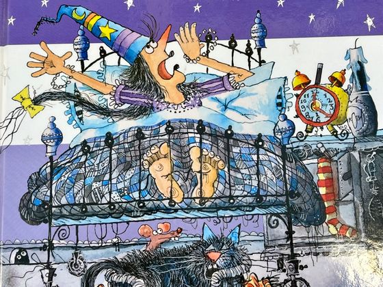 Eine Hexe mit Zauberhut liegt wach im Bett. Davor schläft eine schwarze Katze, der eine Maus über die Schulter schaut.