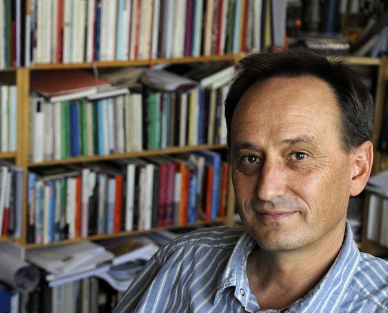 Alois Prinz sitzt, bekleidet mit einem blau-weiß-gestreiften Hemd mit offenem Kragen, vor einem bunten Bücherregal