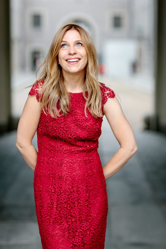 Eine Frau mit langen blonden Haaren und in einem roten Kleid schaut mit einem strahlenden Lächeln nach oben.