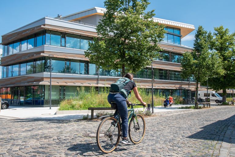 Eine Mitarbeiterin der Gemeindeverwaltung mit dem Fahrrad auf dem Weg zur Arbeit. Im Hintergrund das Rathaus der Gemeinde Gröbenzell.