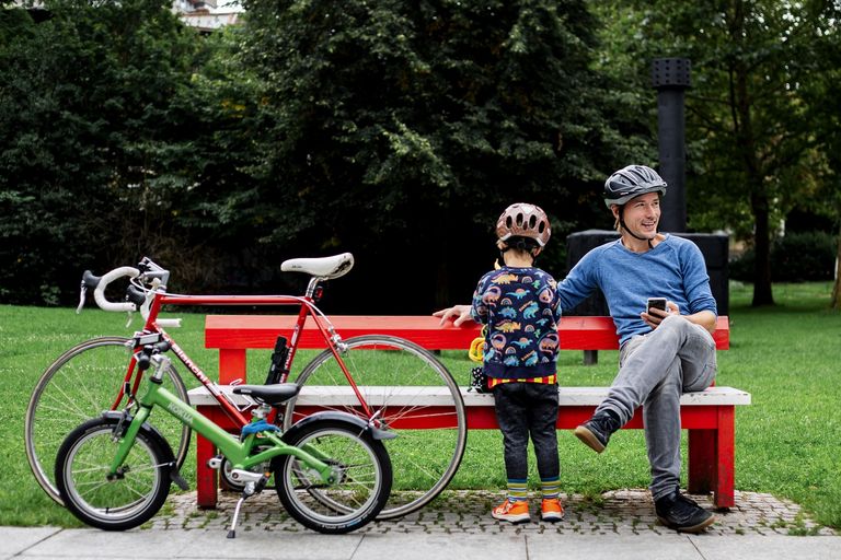 Foto einer roten Bank, auf der ein Mann sitzt mit seinem Sohn (von hinten zu sehen). Davor lehnen 2 Räder.