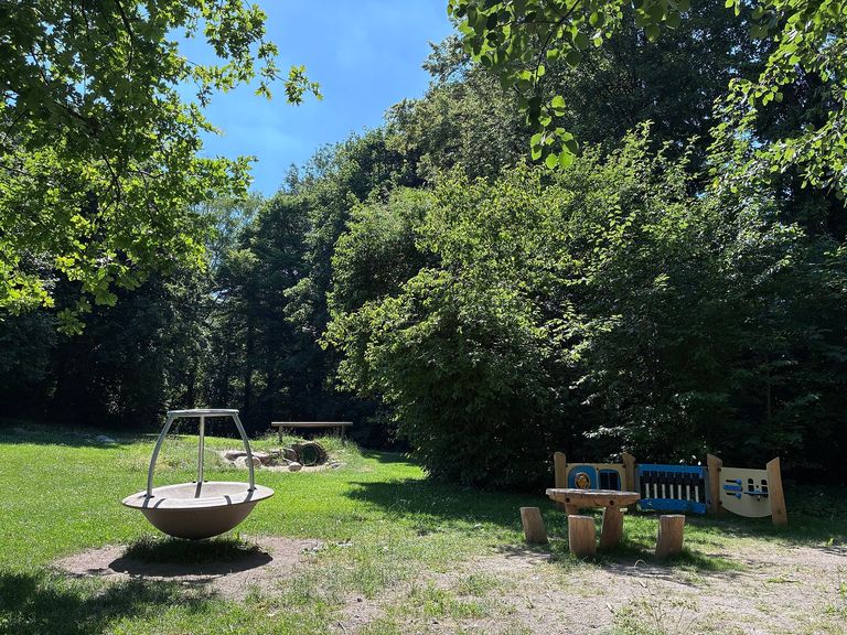 Foto des Spielplatzes mit Spielgeräten, z.B. eine Art Motorik-Bord im Hintergrund, kleiner Tisch mit Hockern, etc.
