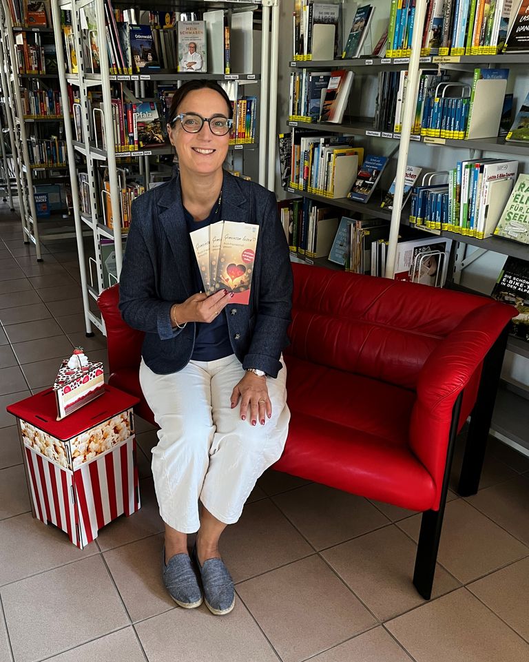 Die Bücherei-Leitung sitzt auf einem roten Sofa in der Bücherei. Im Hintergrund Bücherregale. In den Händen hält sie das Werbematerial zu den "Buch-Freundschaften"